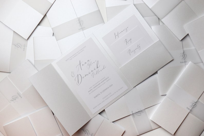May include: A white wedding invitation with a gray envelope. The invitation features the names "Anna & Dominik" in cursive script. The envelope has three pockets labeled "Information", "RSVP", and "Presents".