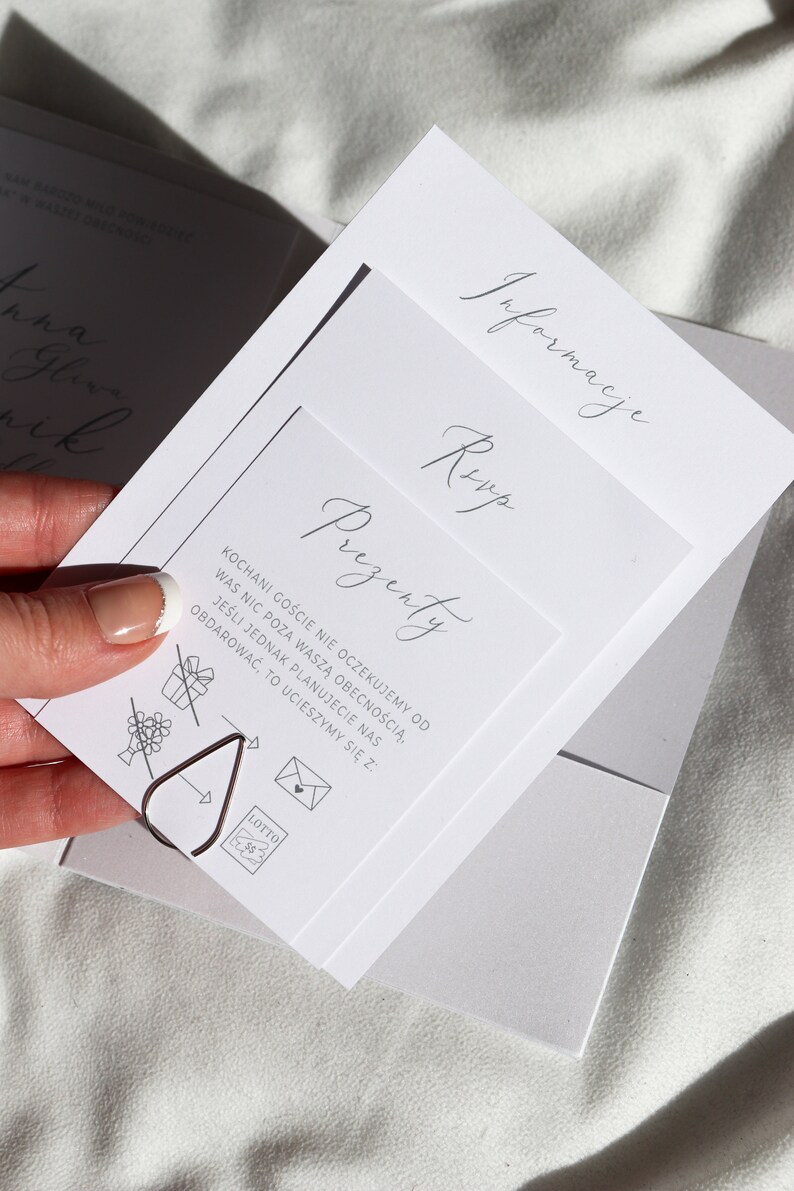 May include: A white card with the text "Prezenty" in cursive script. The card also includes a list of gift ideas, including a bouquet of flowers, a gift box with a ribbon, a letter in an envelope, and a lottery ticket. The text on the card reads: "KOCHANI GOŚCIE NIE OCZEKUJEMY OD WAS NIC POZA WASZĄ OBECNOŚCIĄ. JEŚLI JEDNAK PLANUJECIE NAS ODBDAROWAĆ, TO UCIESZYMY SIĘ Z:"