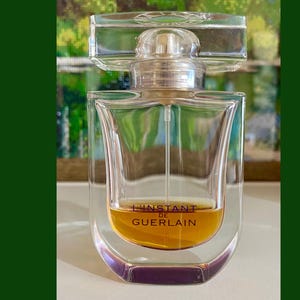 May include: A clear glass perfume bottle with a rectangular shape and a clear, square cap. The bottle contains a golden-coloured liquid and features the text "L'INSTANT DE GUERLAIN". The background is a blurred image of a landscape.