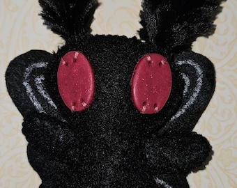 May include: A black plush moth-like creature featuring two large, oval-shaped, glittery dark red eyes and two tall, fuzzy black antennae. The body is covered in soft black fabric with subtle white embroidered wing details on the sides.