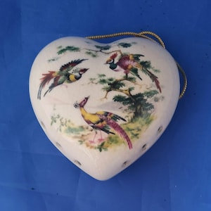 May include: A heart-shaped ceramic ornament with a cream-coloured crackle glaze finish. The surface features a colourful hand-painted scene of three pheasants among green trees and foliage. A thin gold-coloured cord is attached for hanging purposes.