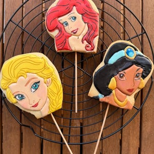 Puede incluir: Tres galletas de azúcar decoradas sobre palitos, con caras de princesas de Disney: Ariel con pelo rojo, Elsa con pelo rubio y Jasmine con pelo negro. Las galletas se exhiben en una rejilla de enfriamiento de alambre negro sobre un fondo de madera.