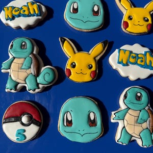 Op de afbeelding: Een verzameling versierde suikerkoekjes met populaire stripfiguren en een naam. De koekjes bevatten Pikachu, Squirtle en een Poké Ball-ontwerp. De naam "Noah" staat op een wolk-vormig koekje.
