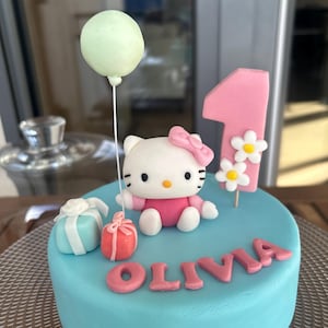 May include: A round, light blue cake decorated for a first birthday. It features a Hello Kitty figure, a pink number one, white daisies, and the name "OLIVIA" in pink letters. A green balloon and two gift boxes complete the festive design.