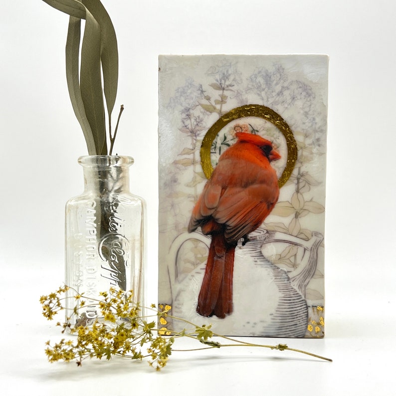 A miniature encaustic painting by Dominique Gustin of a red cardinal saint with a gold halo perched on a white pitcher with gold circles in foreground and muted line drawings of flowers in background