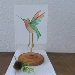 May include: A watercolor painting of a hummingbird in flight, featuring green, orange, and red hues. The artwork is held by a gold-colored metal clip on a wooden base. The base is round and has a decorative edge. The overall theme is nature and art.