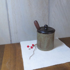 May include: A green and brown ceramic jar with a black lid and a wooden spoon. The jar has a striped pattern and is sitting on a white surface.