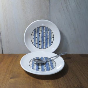 May include: Two white ceramic bowls with a blue and white striped design. The bowls are stacked on top of each other, and a silver spoon is resting on the top bowl.