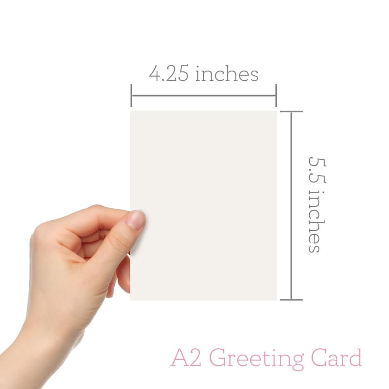 May include: A blank white greeting card measuring 4.25 inches by 5.5 inches. The card is held by a hand in the lower left corner of the image. The text "A2 Greeting Card" is printed in pink below the card.