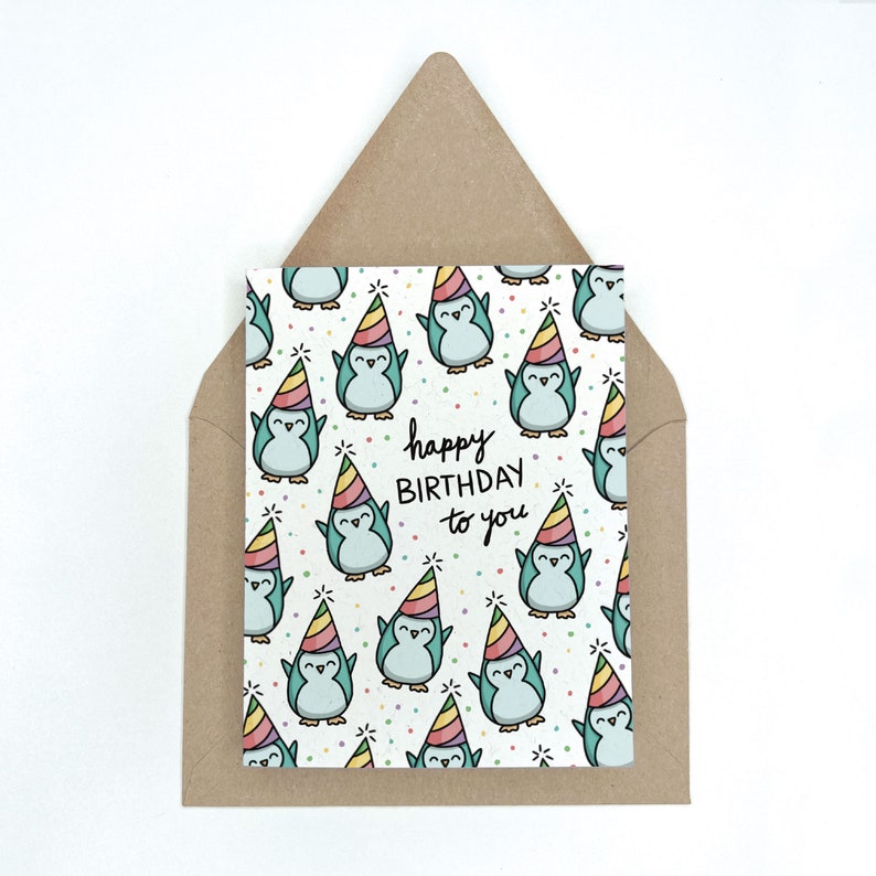 May include: A birthday card featuring a pattern of cartoon penguins wearing party hats. The card has the text "happy BIRTHDAY to you" in black script. The card is inside a brown envelope.