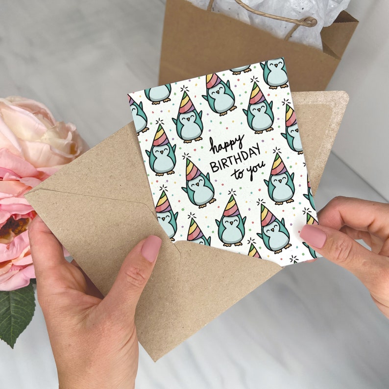 May include: A birthday card with a pattern of cartoon penguins wearing party hats. The card reads "happy BIRTHDAY to you" in black script. The card is inside a brown envelope.