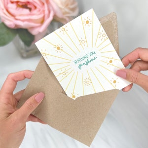 May include: A white card with a yellow sunburst design and the text "Sending you sunshine" in green lettering. The card is being held in a brown envelope.