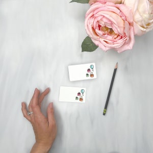 Puede incluir: Dos tarjetas blancas en blanco con un tema de cumplea&ntilde;os. Las tarjetas tienen un globo azul, un regalo rosa y un pastel verde con velas. Las tarjetas est&aacute;n sobre una superficie de m&aacute;rmol blanco.