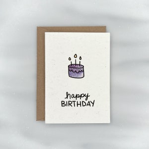 May include: A white card with a purple cake with yellow candles and the text "happy BIRTHDAY" in black.