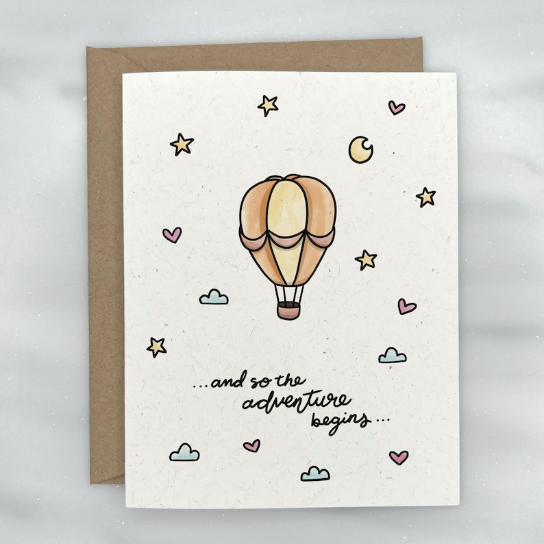 May include: A card with a hand-drawn illustration of a hot air balloon with a light yellow and orange color scheme. The balloon is surrounded by small stars, hearts, and clouds. The text on the card reads "...and so the adventure begins..."