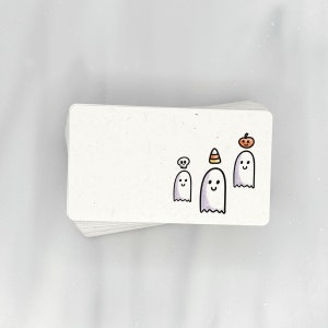 May include: A white card with three ghosts drawn on it. The ghosts have different hats, including a pumpkin, a candy corn, and a skull.
