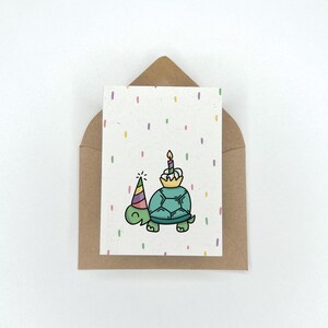 May include: A birthday card with a green turtle wearing a party hat and holding a cupcake with a lit candle. The card has a white background with colorful confetti.