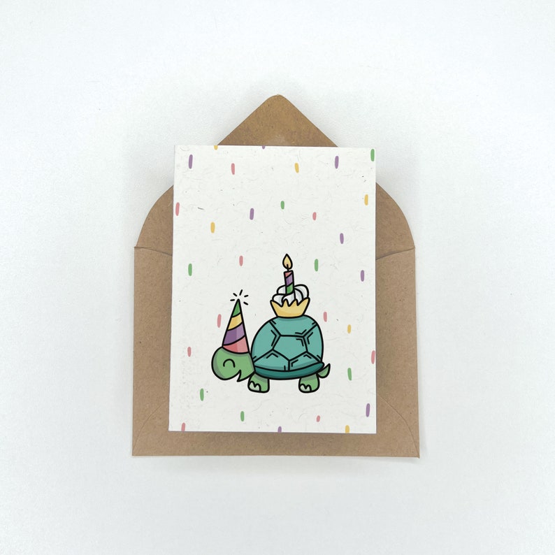 May include: A birthday card with a green turtle wearing a party hat and holding a cupcake with a lit candle. The card has a white background with colorful confetti.