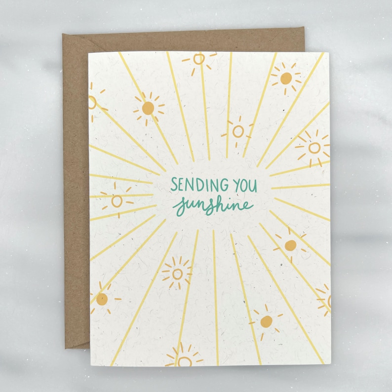 May include: A white card with a yellow sunburst design and the text "Sending you sunshine" in green lettering.