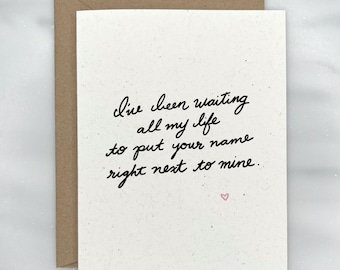 May include: A white card with a handwritten message that reads "I've been waiting all my life to put your name right next to mine." A small pink heart is drawn at the bottom of the card.