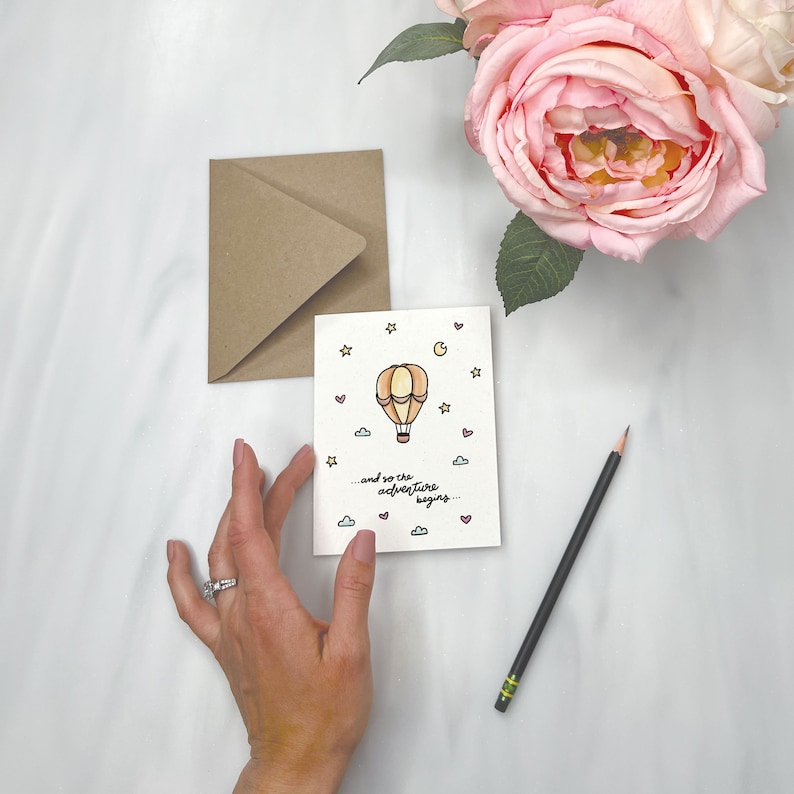 May include: A white card with a hand-drawn illustration of a hot air balloon with a basket and a crescent moon. The balloon is orange and yellow with pink and blue accents. The card has the text "...and so the adventure begins..." in black cursive writing.