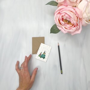 May include: A white card with a colorful illustration of a turtle wearing a party hat and holding a cake. The card is on a white surface with a brown envelope and a black pencil.