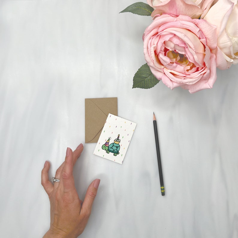May include: A white card with a colorful illustration of a turtle wearing a party hat and holding a cake. The card is on a white surface with a brown envelope and a black pencil.