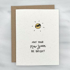May include: A white card with a brown envelope. The card has a simple drawing of a sun with sunglasses and the text "May your New Year be bright".