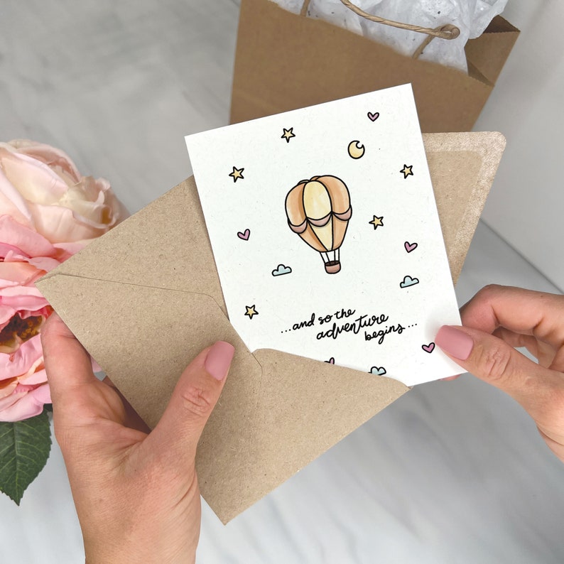 May include: A card with a hand-drawn illustration of a hot air balloon with a pink, yellow, and orange color scheme. The card has the text "...and so the adventure begins..." in black cursive writing.