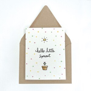 Peut inclure: Une carte blanche avec un motif de pois verts, jaunes et roses. La carte a un soleil avec un visage souriant, une plante en pot avec un c&oelig;ur et le texte "hello little sprout".