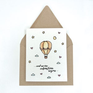 May include: A card with a hand-drawn illustration of a hot air balloon floating in the sky with stars, a crescent moon, and clouds. The text on the card reads "...and so the adventure begins..."