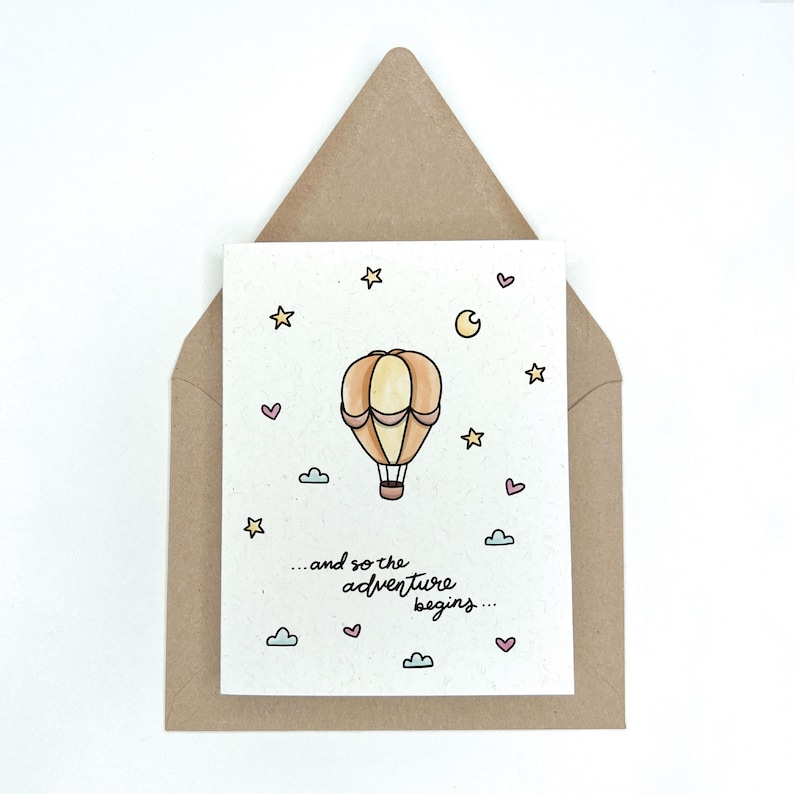 May include: A card with a hand-drawn illustration of a hot air balloon floating in the sky with stars, a crescent moon, and clouds. The text on the card reads "...and so the adventure begins..."