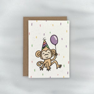 May include: A birthday card featuring a cartoon monkey wearing a colorful party hat and holding a purple balloon. The card has a white background with confetti.