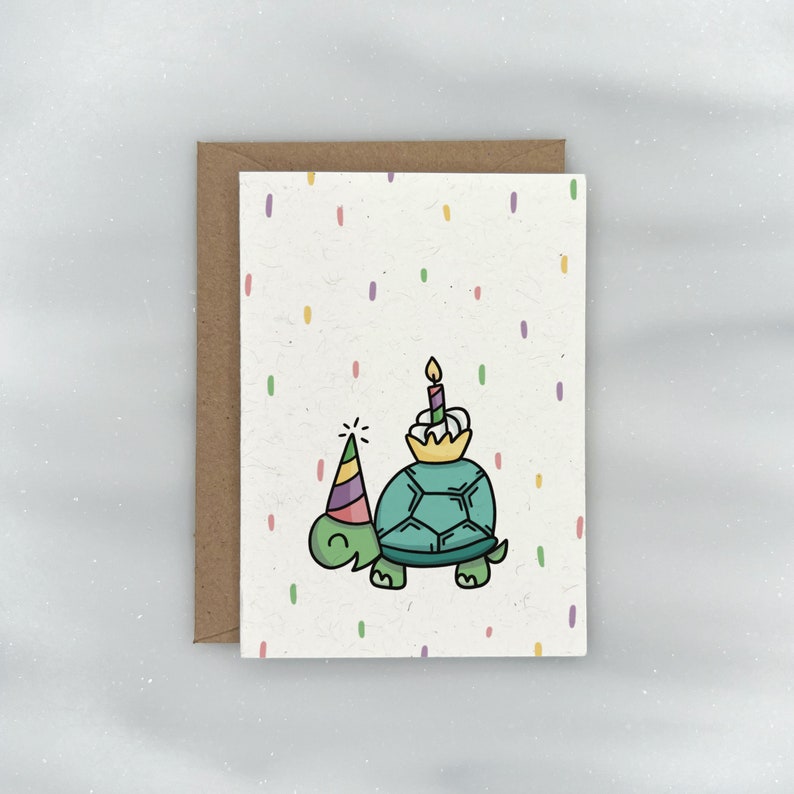 May include: A birthday card with a green turtle wearing a party hat and carrying a cupcake with a lit candle on its back. The background is white with colorful confetti.