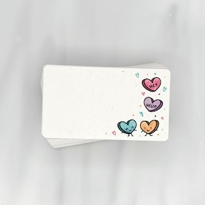 May include: A stack of white note cards with a colorful heart design on the top card. The hearts have faces and say "Hey", "Hello", and "Love".