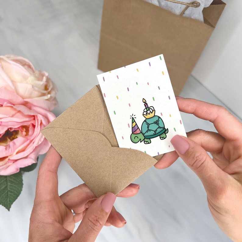 May include: A brown envelope with a white card inside. The card has a colorful confetti background and a cartoon illustration of a turtle wearing a party hat and holding a birthday cake.
