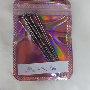 May include: A package of silver-colored hair pins in a pink iridescent pouch. The pouch has a zip closure and a small label with the numbers "425 36" written on it. The background is a soft, white, fluffy surface.