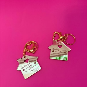 Puede incluir: Dos llaveros con forma de casa con herrajes dorados y una superficie reflectante. Cada llavero tiene un recorte en forma de corazón y texto grabado. Los llaveros están sobre un fondo rosa brillante. El texto en los llaveros dice "Qualunque cosa accada, ci vediamo a casa G&R."