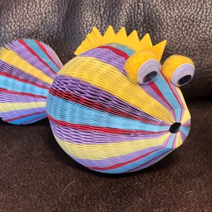 May include: A colorful paper fish with yellow, red, blue, and purple stripes. The fish has large googly eyes and a yellow crown on its head.