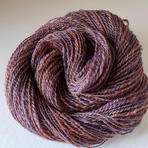 May include: A ball of yarn in shades of purple, brown, and yellow. The yarn is tightly wound, with visible strands and a textured appearance. Ideal for knitting or crocheting projects.