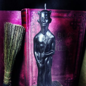 May include: A black candle in the shape of a devil figure, holding a trident. The candle is positioned in front of a stack of antique books with red covers and a bundle of dried grass.