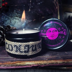 Könnte beinhalten: Ein schwarzes Kerzengefäß mit einem Jute-Etikett, auf dem "LONE WOLF" steht. Die Kerze ist angezündet und das Gefäß steht auf einem alten Buch. Der Deckel des Gefäßes hat ein pinkfarbenes und schwarzes Design mit den Worten "Wicked World" und einem Mondgesicht.