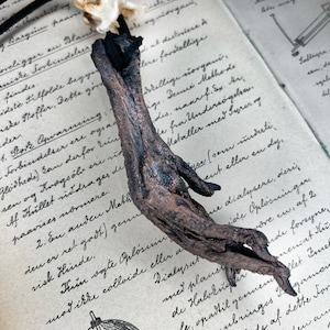 May include: A dark brown, gnarled, wooden pendant with a black cord. The pendant is shaped like a hand with long, slender fingers. The pendant is hanging over an open book with handwritten text.