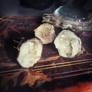 May include: Three geode rings with silver-colored metal bands and edges. The geodes have a crystalline interior and are set on a dark, textured surface. A clear glass turtle figurine is in the background.