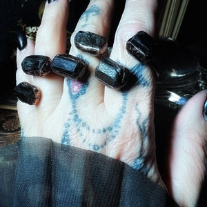May include: Close-up of several dark, rectangular crystals resting on a hand with tattoos. The crystals are a deep, opaque black with some showing a hint of a lighter color. The nails are painted black, and a dark fabric is visible.
