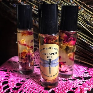 May include: Three glass bottles with black roll-on tops filled with a clear liquid and rose petals. The label on the center bottle reads "Apothecary of Curiosities Love Spell Oil" with a dragonfly illustration.