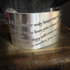 Könnte beinhalten: Ein breiter Armreif aus silberfarbenem Metall mit einer Gravur des Liedtextes aus dem Song Dreams von Fleetwood Mac. Der Text lautet: Oh, thunder only happens when it's raining. Players only love you when they're playing. Women, they will come and they will go. When the rain washes you clean, you'll know.