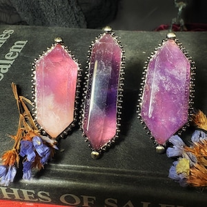 May include: Three gemstone pendants with silver-colored metal trim. The crystals are various shades of purple and pink. The pendants are arranged on a dark surface with the text "HES OF SALEM" visible. Dried flowers are in the foreground.