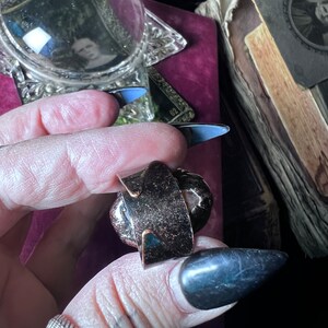 May include: A dark metallic adjustable ring featuring a wide, flat band with a textured, speckled surface. The ring has an open-back design, allowing for size adjustments, and displays a deep, bronze-toned finish with subtle light reflections.