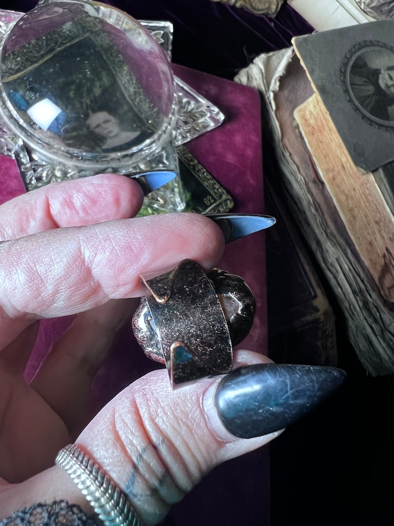 May include: A dark metallic adjustable ring featuring a wide, flat band with a textured, speckled surface. The ring has an open-back design, allowing for size adjustments, and displays a deep, bronze-toned finish with subtle light reflections.